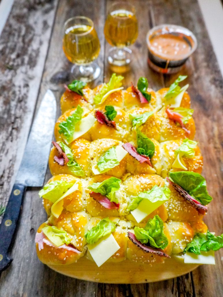 Une brioche garnie de charcuterie, de tranches de fromage et de salade, posée sur une planche à découper